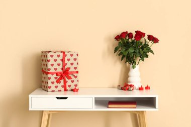 White desk with bouquet of roses, gift box and candles near beige wall. Valentine's Day celebration
