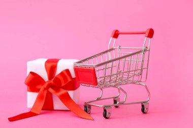 Pembe arka planda hediye kutusu olan küçük bir alışveriş arabası. Sevgililer Günü kutlaması