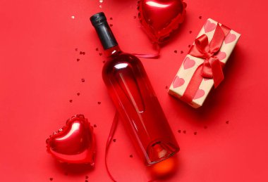 Hediyeli bir şişe şarap, kırmızı arka planda balonlar ve konfeti. Sevgililer Günü kutlaması