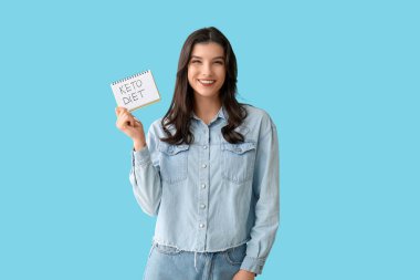 Elinde mavi arka planda KETO diyeti yazılı not defteri olan mutlu, genç bir kadın.