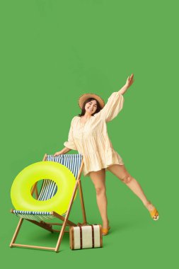 Yeşil arka planda şişirilebilir yüzüğü, bavulu ve şezlonguyla güzel genç bir kadın.