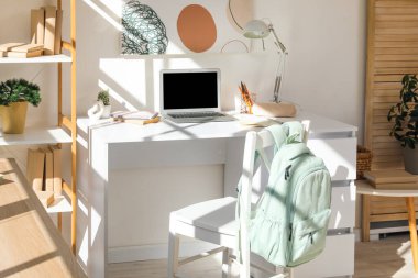 Kırtasiyeli, sırt çantalı ve laptoplu modern okul masası beyaz duvarın yanında.