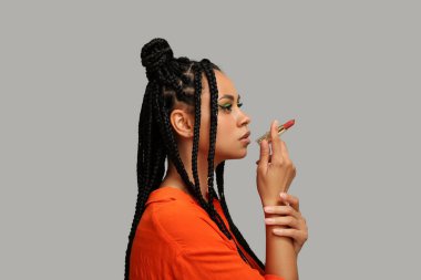 Saçları örgülü ve makyajlı genç bir Afro-Amerikalı kadın gri arka planda ruj tutuyor.