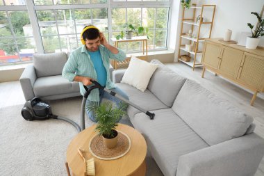 Genç adam kanepeyi süpürüyor ve odada müzik dinliyor.