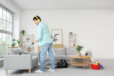 Genç adam kanepeyi süpürüyor ve odada müzik dinliyor.