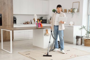 Bilgisayarlı genç adam mutfakta halı süpürüyor.