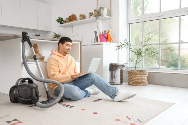 Dizüstü bilgisayarı ve elektrikli süpürgesi olan genç bir adam mutfakta.