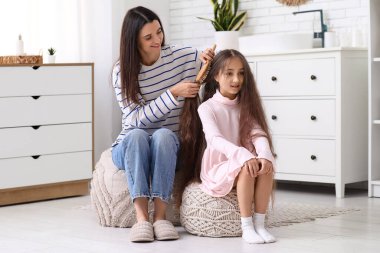 Genç anne banyoda puf üzerinde otururken küçük kızının saçlarını tarıyor.