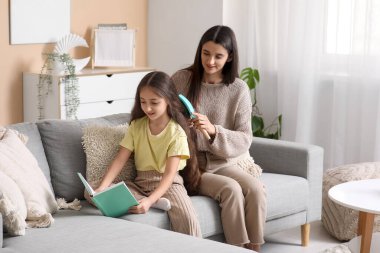 Genç anne, evdeki kanepede oturup kitap okuyan küçük kızının saçını tarıyor.