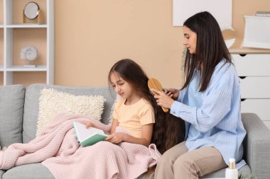 Genç anne, evdeki kanepede oturup kitap okuyan küçük kızının saçını tarıyor.