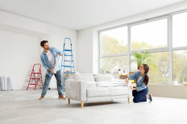 Genç bir çift, yeni evlerinde tamir sırasında streç filmle kanepeyi sarıyorlar.