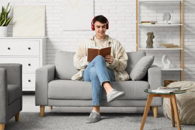Evdeki kanepede kitap okuyan kulaklıklı yakışıklı genç adam.