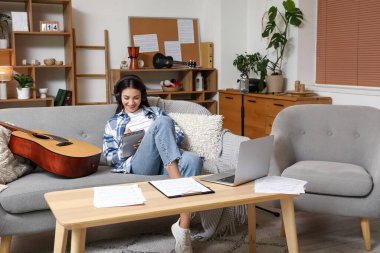 Evdeki kanepede dizüstü bilgisayarı, kulaklığı ve gitar yazma notları olan genç ve güzel bir besteci.