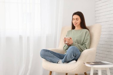 Elinde çay bardağıyla evdeki ışık perdesinin yanındaki koltukta oturan genç kadın.