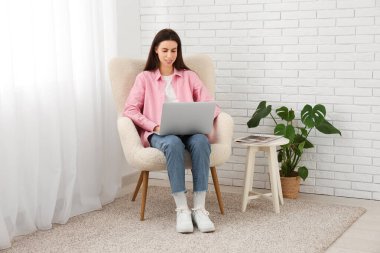 Evdeki ışık perdesinin yanındaki koltukta dizüstü bilgisayar kullanan genç bir kadın.