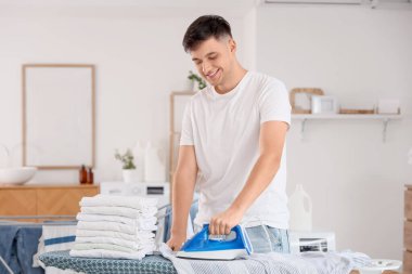 Yakışıklı genç adam evde giysileri ütülüyor.