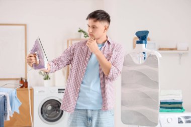 Yakışıklı, genç, düşünceli bir adam evdeki çamaşır odasında demir ve vapur arasında seçim yapıyor.