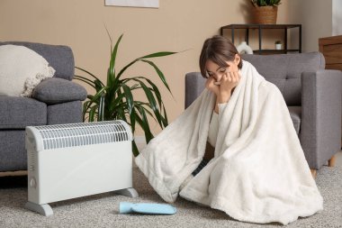 Battaniyeli ve sıcak su şişeli genç bir kadın ısıtıcının yanında ısınıyor.