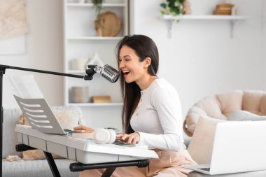 Evdeki kanepede mikrofonlu genç bir kadın sentezleyici çalıyor.