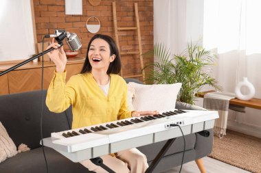 Evdeki kanepede mikrofonlu genç bir kadın sentezleyici çalıyor.