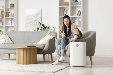 Elinde çay ve hava temizleyicisiyle evdeki koltukta oturan genç bir kadın.