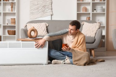 Evde elektrikli konteyner ısıtıcısının yanında oturan ekoseli çay bardağıyla genç bir adam.