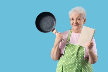 Önlüklü, kızarmış tavalı ve mavi arka planda yemek tarifi kitabı olan yaşlı bir kadın.