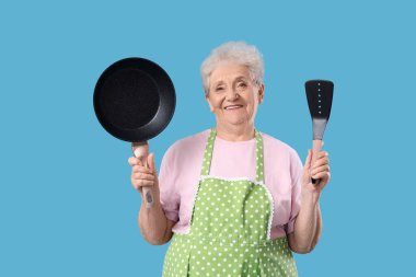 Önlüklü yaşlı kadın, mavi arka planda kızartma tavası ve spatulası var.