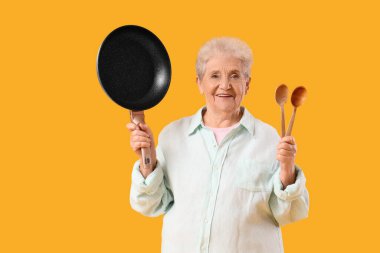 Kızartma tavası ve sarı arka planda kaşıkları olan yaşlı bir kadın.
