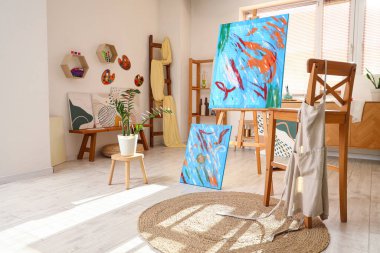 Sanatçı atölyesinin içinde resimler, ev bitkisi ve önlük var.