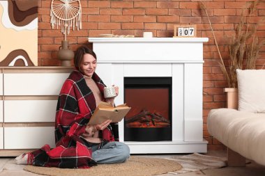 Güzel, genç, mutlu bir kadın, elinde kakao bardağıyla şöminenin yanında kitap okuyor.