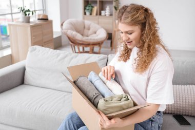 Evdeki kanepede yeni kıyafetlerle paket kutusunu açan genç bir kadın.