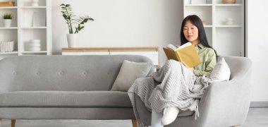 Genç Asyalı kadın evdeki kanepede otururken kitap okuyor.