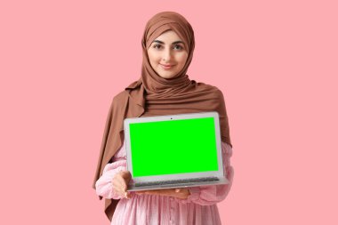 Müslüman kadın pembe arka planda yeşil ekranlı modern dizüstü bilgisayar tutuyor