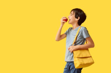 Çantalı küçük çocuk sarı arka planda elma yiyor.