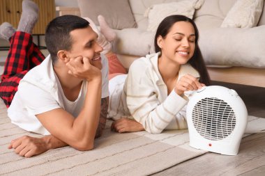 Evde elektrikli fan ısıtıcısının yanında ısınan genç bir çift var.