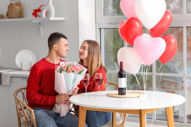 Kalp şeklinde balonları, bir buket çiçekleri ve evde sevgililer gününü kutlayan şarap kadehleri olan genç ve güzel bir çift.