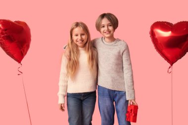 Küçük çocuklar, hediye ve kalp şeklinde balonlarla pembe arka planda sarılıyorlar. Sevgililer Günü kutlaması