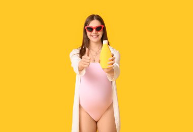 Güneş kremi şişesiyle mayolu hamile bir kadın sarı arka planda baş parmak hareketini gösteriyor.