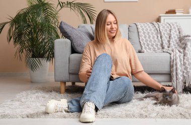 Orta yaşlı bir kadın evde kedisiyle yerde oturuyor.