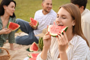 Genç bir kadın arkadaşlarıyla parkta piknik yaparken karpuz yiyor.