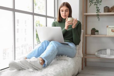 Evde bir fincan sıcak çay ve dizüstü bilgisayarı olan genç bir kadın.