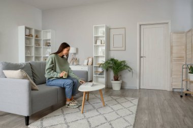 Gri koltukta kahve fincanı olan genç bir kadın oturma odasında dizüstü bilgisayar kullanıyor.