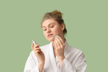 Yeşil arka planda fondöten şişesi ve makyaj süngeri olan güzel bir genç kadın.