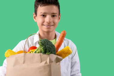 Yeşil arka planda farklı sebzelerle dolu kese kağıdı tutan sevimli küçük çocuk.
