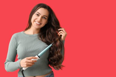 Beautiful young woman with curling iron on red background