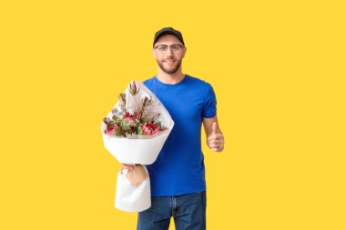 Elinde bir buket güzel çiçekle genç teslimatçı sarı arka planda baş parmak hareketi gösteriyor.