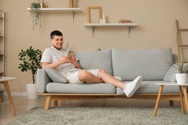 Oturma odasındaki koltukta oturan cep telefonlu genç adam.