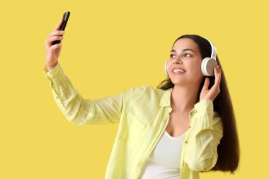 Güzel, genç, mutlu, kulaklıklı bir kadın müzik dinliyor ve sarı arka planda selfie çekiyor.
