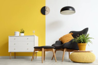 Şık oturma odasının içinde siyah koltuk, çekmeceler, lamba ve saat sarı duvarda.